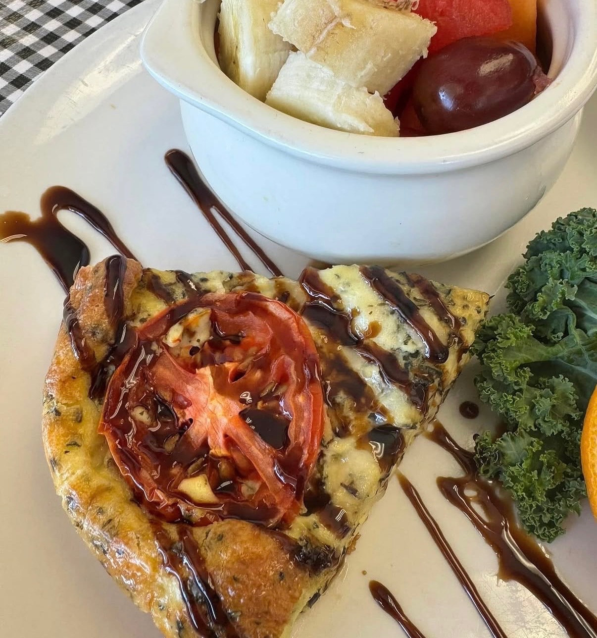 A slice of quiche topped with roasted tomato and balsamic drizzle, served with fresh fruit